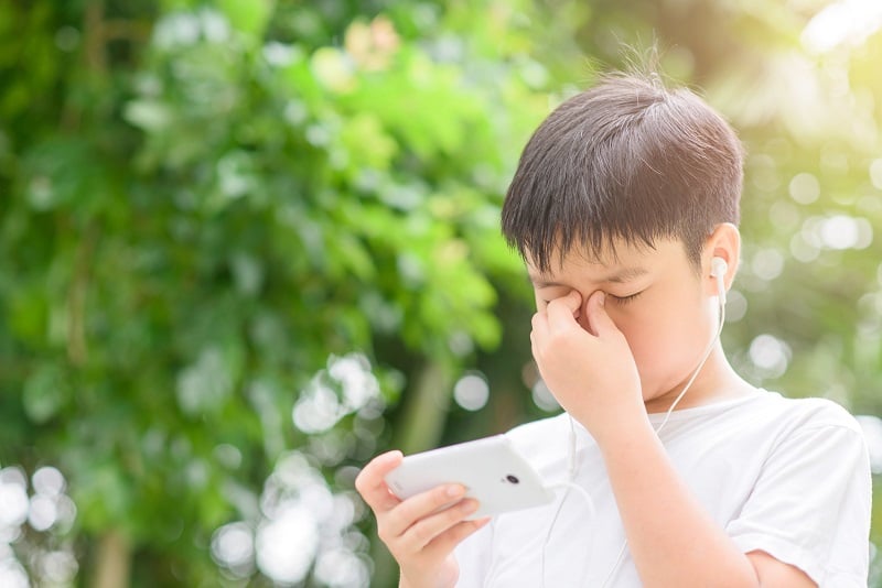 Young Asian Thai boy using a smartphone to play game and listen to music without care the surrounding and he has a problem with headache and eye pain.